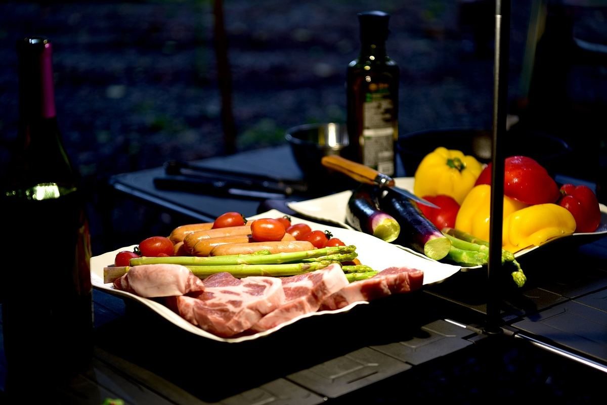 Assiette de charcuterie et légumes frais sur table de camping en soirée, avec bouteilles de vin et fruits à proximité Assiette de charcuterie et légumes frais sur table de camping en soirée, avec bouteilles de vin et fruits à proximité